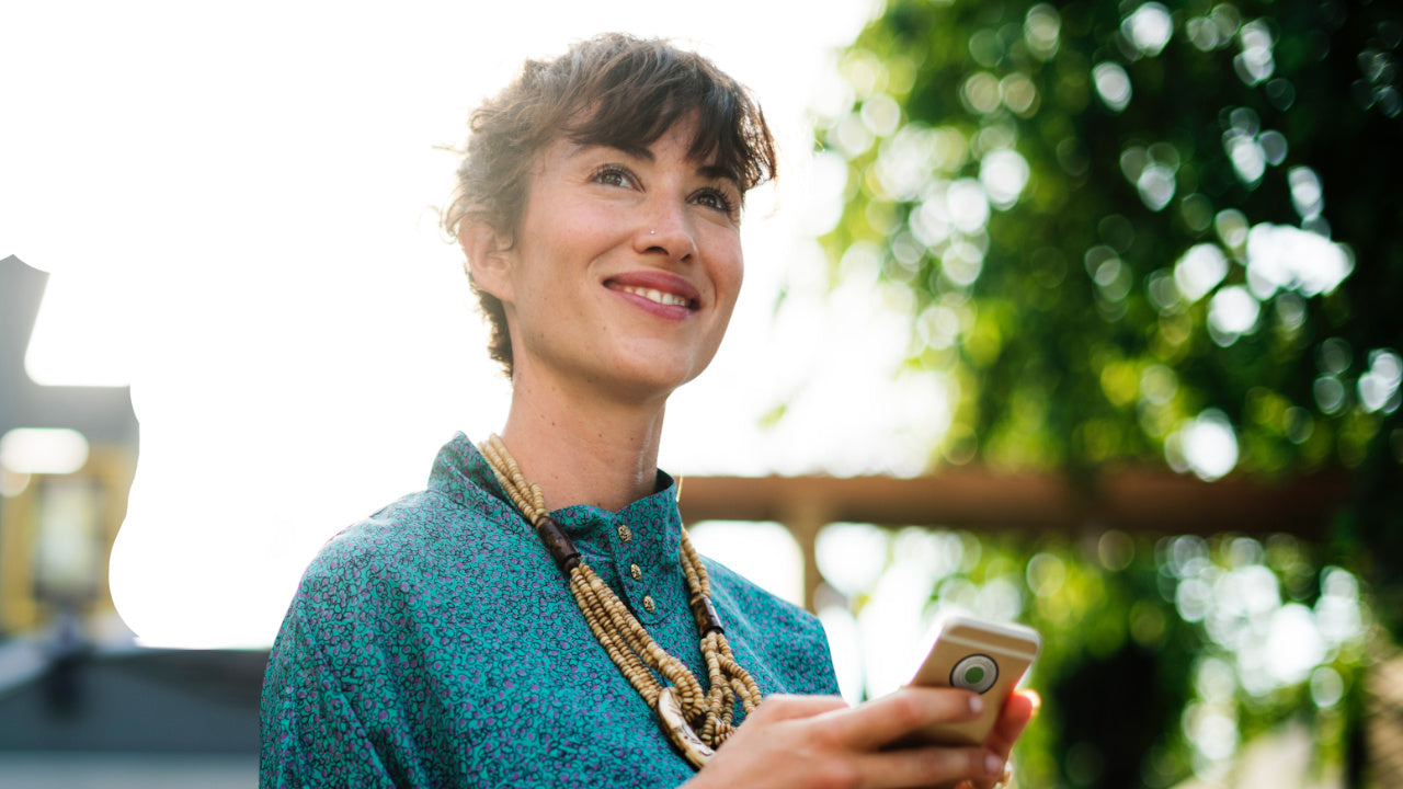 Eine lächelnde braunhaarige Frau, die ein Handy mit einem Vita Chip 2.0 in der Hand hält, mit einem verschwommenen Hintergrund mit einem hellweißen Himmel und etwas Grünzeug.